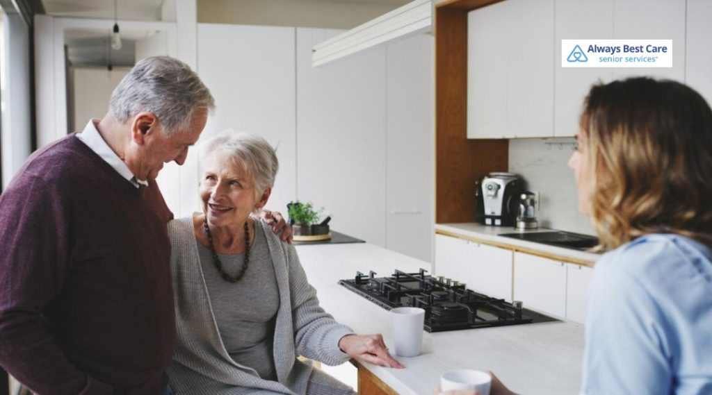 Smiling older couple chats with a caregiver in a bright kitchen over coffee.
