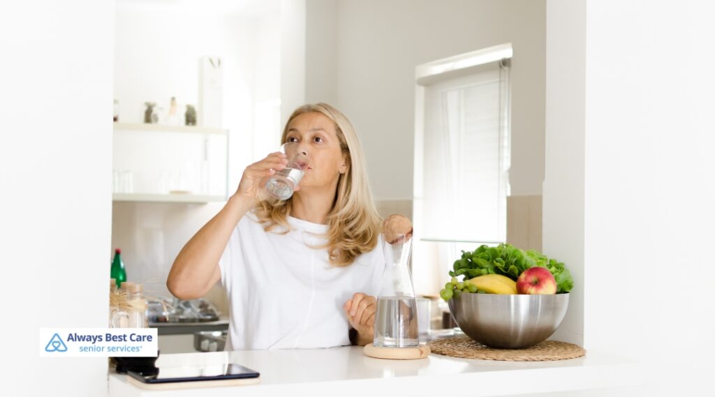 Woman drinking water at a kitchen counter beside a water carafe and a bowl of fresh produce.