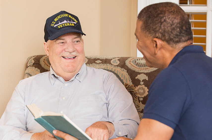 SENIOR VETERAN SMILING WITH A CAREGIVER WHILE READING BOOK