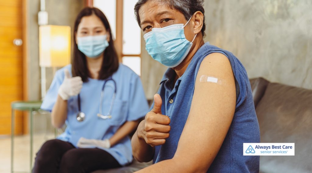 A senior man receiving a vaccine shot, accompanied by a healthcare worker in a medical mask and gloves, giving a thumbs-up after the injection.