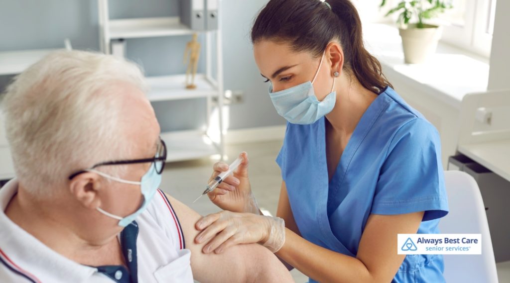 A senior woman in a brown top, wearing glasses and a face mask, receiving a vaccine shot from a healthcare provider in a white coat and blue gloves.