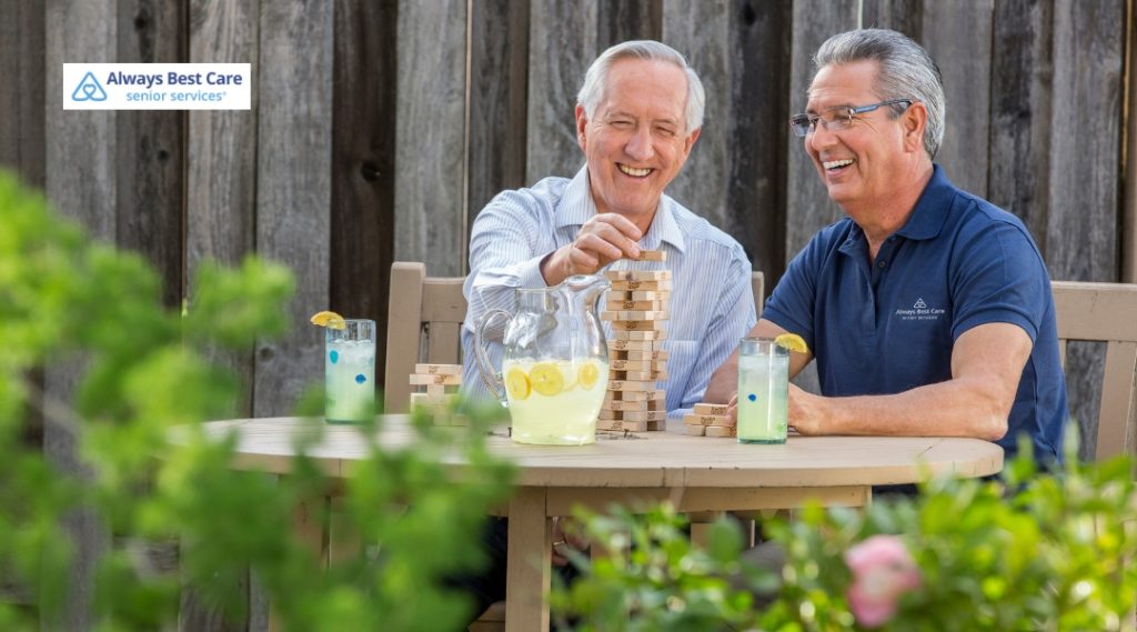 Two men, one in a blue shirt and the other in a grey shirt, are enjoying a game of Jenga on a wooden table in a garden. They are both smiling, and there are glasses of lemonade on the table, with a green background of plants and a wooden fence.