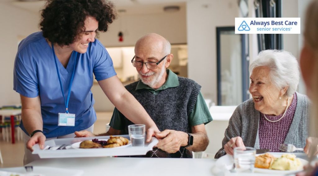 Caregiver serving meals and supporting older adults during assisted living dining activities.