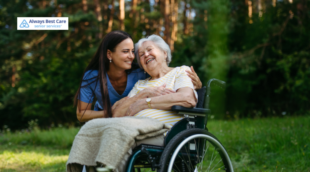 Caregiver embracing a laughing older woman in a wheelchair outdoors.