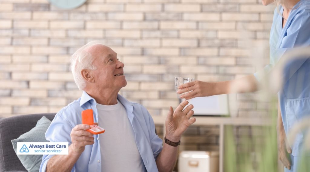 Smiling older man sitting on a couch, holding a pill organizer as a caregiver in scrubs hands him a glass of water.