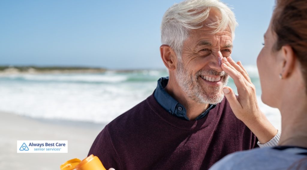 An older man smiling on the beach while a caregiver applies sunscreen on his nose, enjoying the sun together.