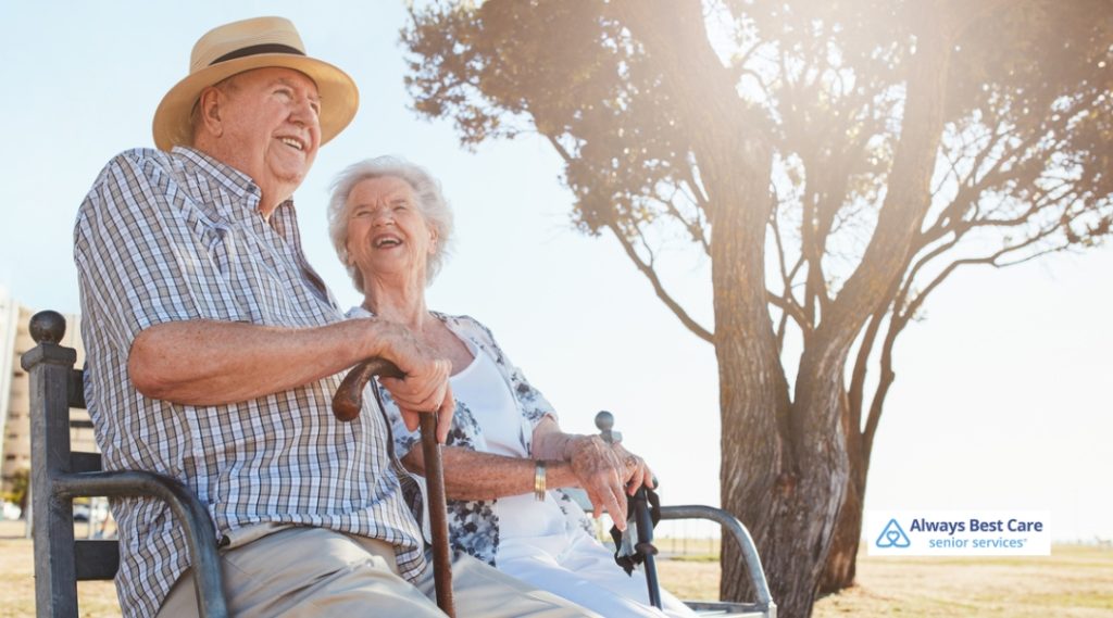 A senior couple sitting on a bench in the park, smiling and enjoying each other's company while the sun shines behind them.