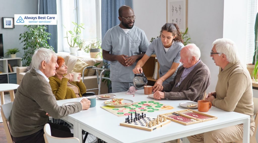 A group of seniors playing board games together with the assistance of two caregivers. One caregiver is pouring tea while others are seated at a table enjoying snacks and drinks. The image includes the "Always Best Care" logo in the top left corner.