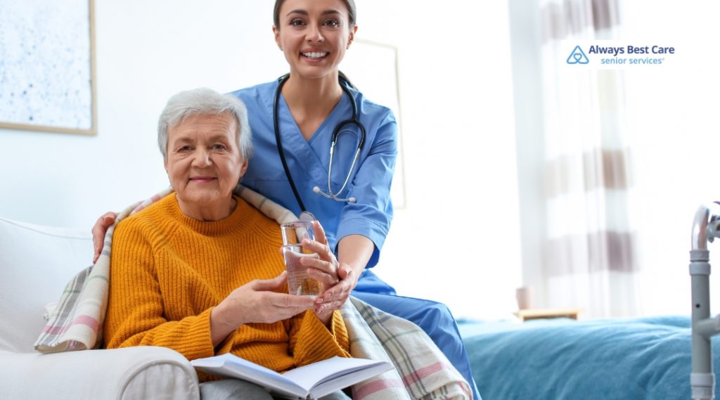 A caregiver in a blue medical uniform smiling and offering a glass of water to an elderly woman wearing a cozy yellow sweater, sitting comfortably in a home setting. The "Always Best Care" logo is in the top left corner.