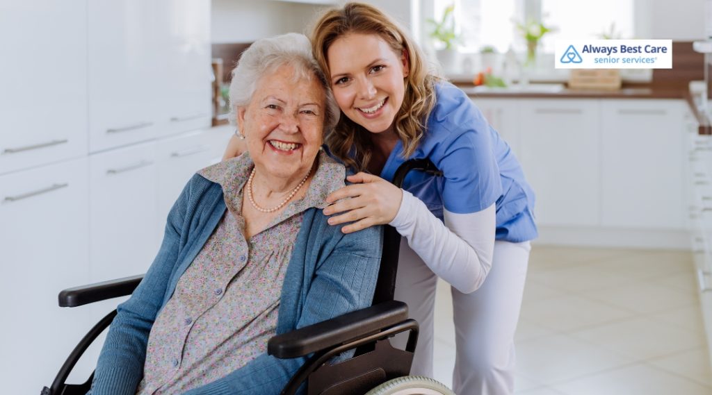 A caregiver in a blue medical uniform smiling beside an elderly woman in a wheelchair, both looking happy and comfortable in a bright kitchen. The "Always Best Care" logo is in the top left corner.