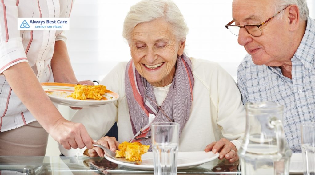 Smiling senior couple receives a hot plate of casserole from a caregiver, capturing the joy of mealtime support.
