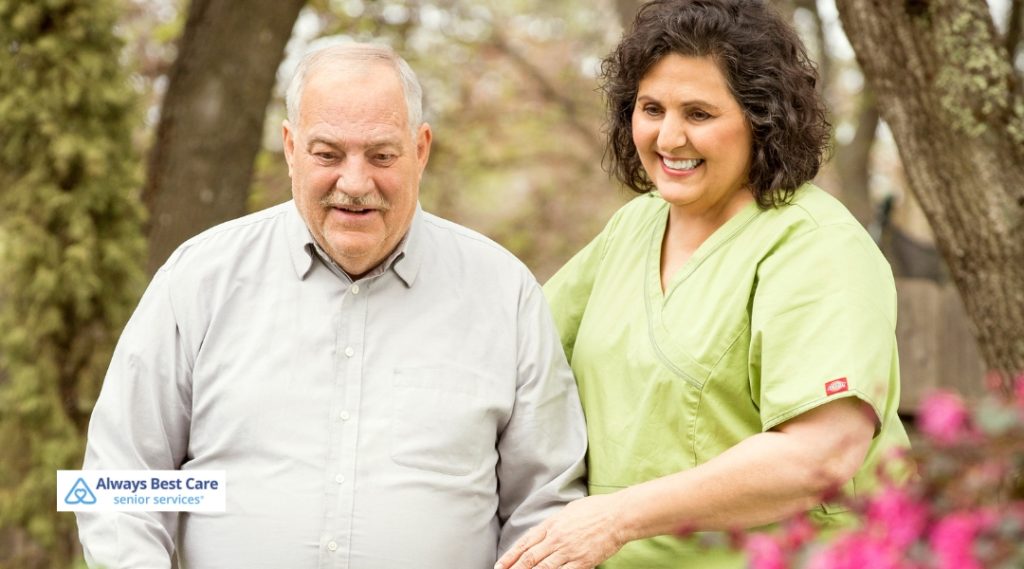 A female caregiver in a green scrub top walks outside with an elderly man, offering assistance and companionship. The Always Best Care logo appears on the image.