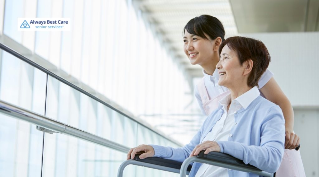 A caregiver in white scrubs helps an elderly woman walk with a walker in a well-lit, modern building with large windows. The caregiver smiles at the woman, providing support and guidance as they move through the space.