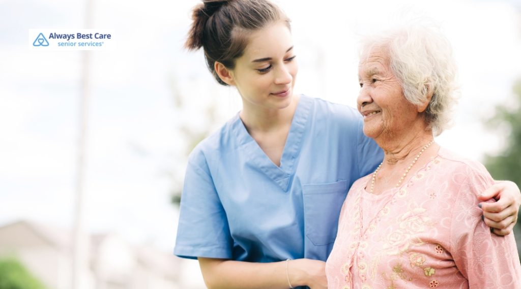 A caregiver in scrubs supports an elderly woman walking outside in a residential area. The woman smiles as she walks with the help of her caregiver, who stands beside her with a reassuring expression, highlighting a positive outdoor care experience.
