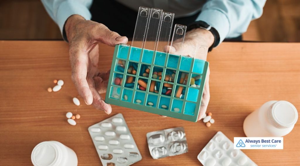 An older adult’s hands hold a blue weekly pill organizer filled with various prescription and over-the-counter medications, with additional pill bottles and blister packs scattered across a wooden table.
