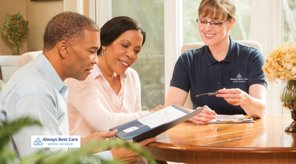 A care coordinator in a navy Always Best Care polo shirt presents documents to a middle-aged couple, seated together at a dining table in a well-lit home. The logo is included at the bottom left.
