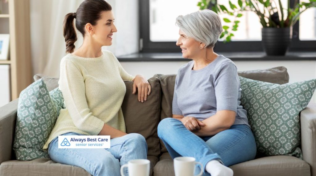 A caregiver and an elderly woman sitting comfortably on a couch, chatting and enjoying a cup of coffee together. The image emphasizes the bond between caregivers and seniors in a relaxed home setting.