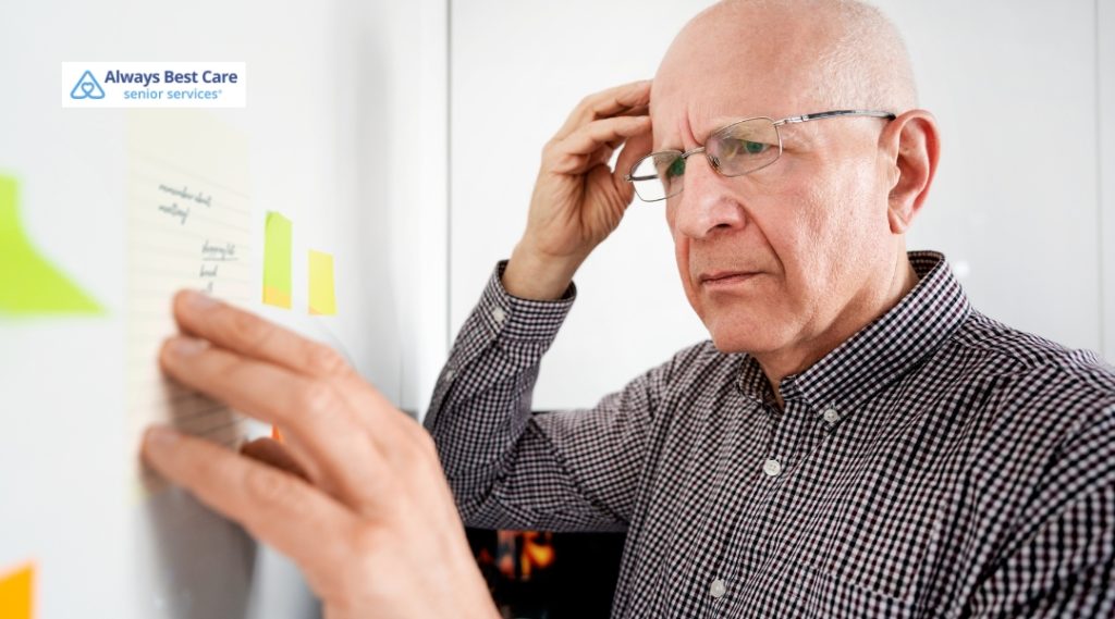 An elderly man with glasses and a checkered shirt looks thoughtfully at a note he is holding against a wall covered with sticky notes. His expression shows concentration as he scratches his head.