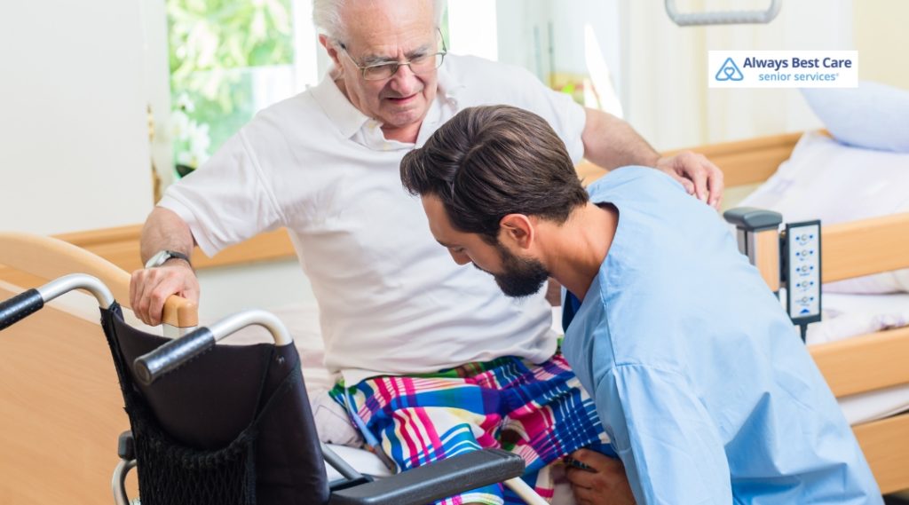 A male caregiver in blue scrubs is kneeling beside an elderly man who is sitting on the edge of a bed. The elderly man, wearing glasses and a white shirt, is holding onto the bed frame and looking down toward a wheelchair positioned in front of him. The room is softly lit with a window showing greenery outside. The Always Best Care logo appears in the upper right corner.