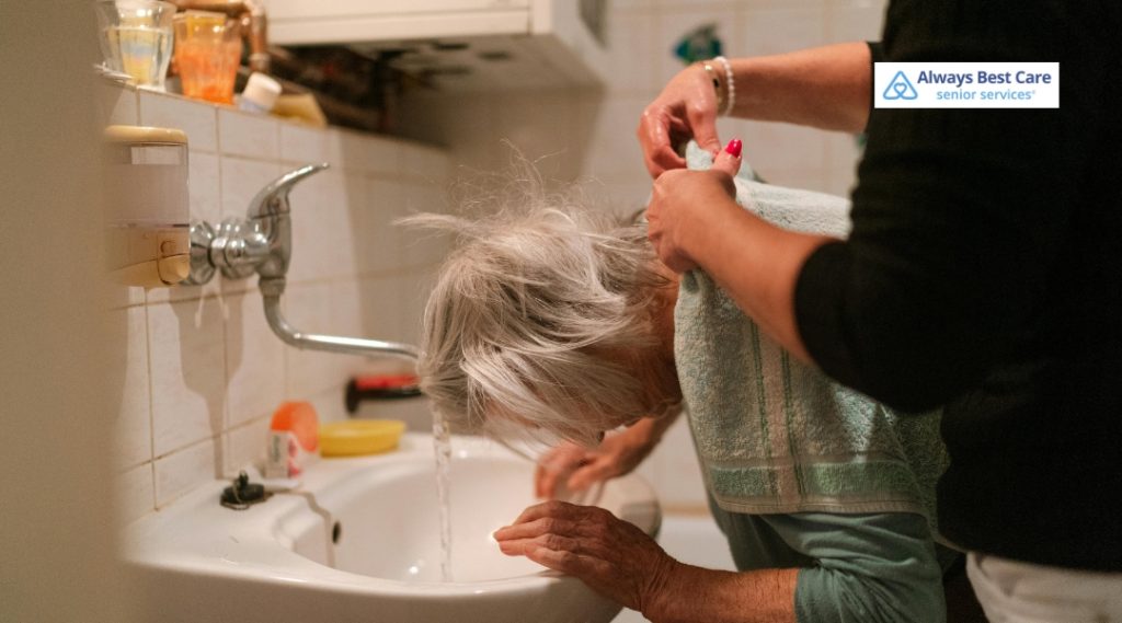 A caregiver is gently helping an elderly woman with gray hair wash her face at a sink. The caregiver is holding a towel around the woman’s shoulders as the woman leans over the sink under running water. The scene takes place in a modest bathroom. The Always Best Care logo is in the upper right corner.
