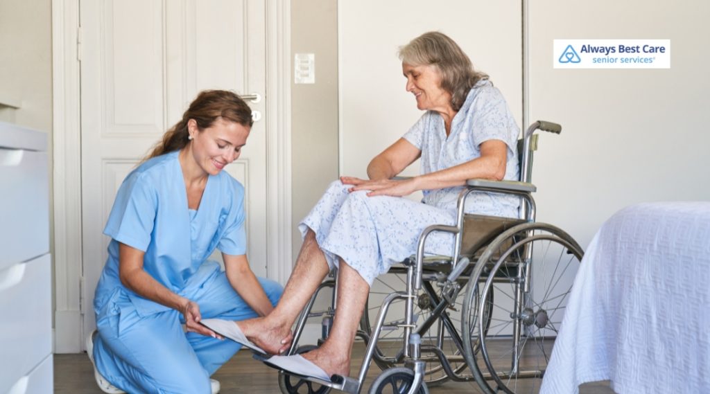A young female caregiver in light blue scrubs is assisting an elderly woman sitting in a wheelchair. The elderly woman, wearing a patterned dress, smiles down at her feet as the caregiver adjusts or places a protective foot covering. The room looks like a cozy living or care space with soft lighting. The Always Best Care logo is positioned in the upper right corner.