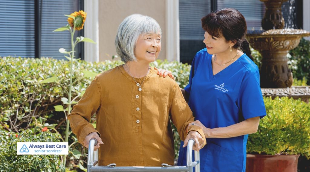 Caregiver assisting a senior woman with a walker while walking in a garden, with both smiling and enjoying the fresh air.