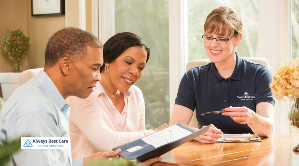 A caregiver is assisting a senior couple while reviewing a document in a well-lit living room. The caregiver is showing them something, and both seniors are attentive. The Always Best Care logo is in the top left corner.