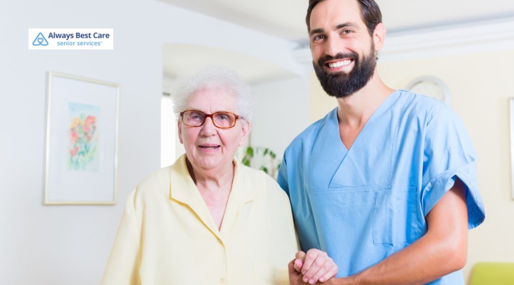 An elderly woman with gray hair and glasses stands next to a male caregiver in a blue uniform. They are both smiling, with the caregiver holding her hand. The background features a light and airy room with framed artwork on the walls.