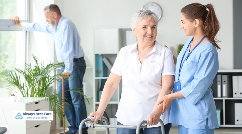 A senior woman with short gray hair uses a walker while being assisted by a female caregiver in a blue uniform. The caregiver is helping her walk, and a man with a beard stands by the window in the background. The room is well-lit with plants and office supplies.