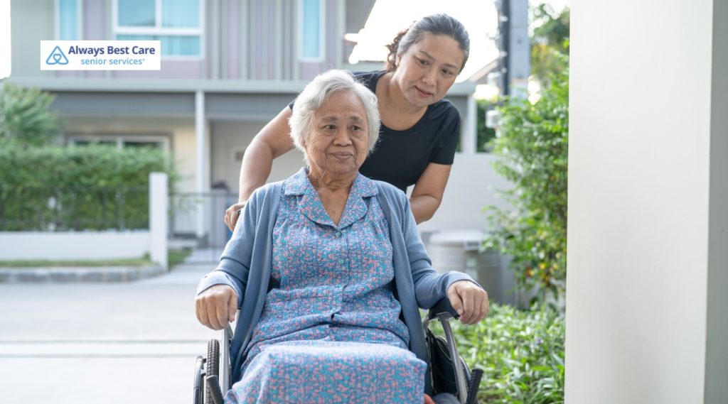 An elderly woman in a wheelchair is outside, being pushed by a younger woman wearing a black shirt. They appear to be in a residential area with a modern building in the background. The elderly woman looks content, and the younger woman is attentively guiding her. The "Always Best Care senior services" logo is in the top left corner.