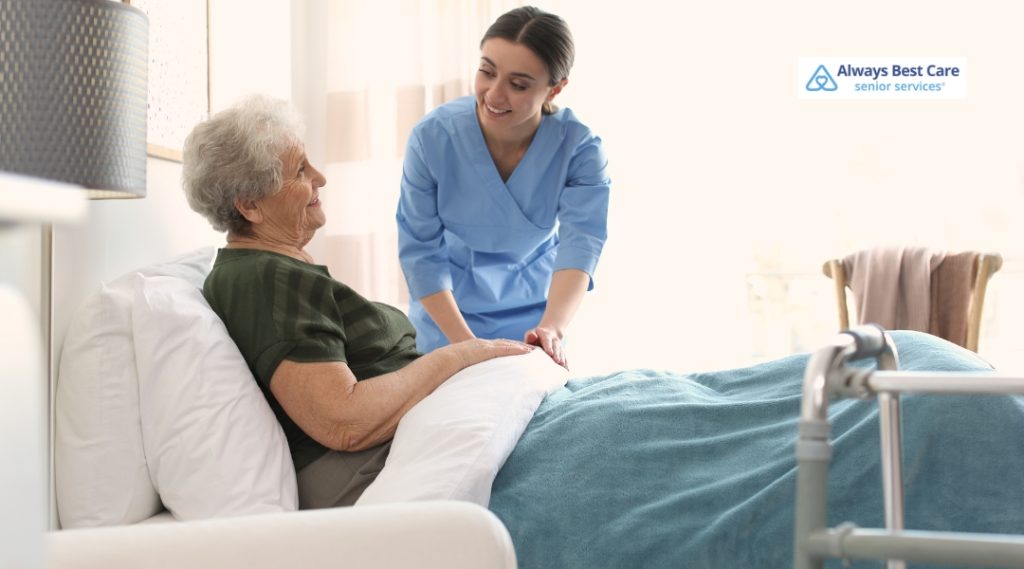 A young female nurse in blue scrubs is warmly engaging with an elderly woman resting in bed. The elderly woman is smiling and looks comfortable, covered with a teal blanket. The scene reflects compassionate bedside care with the "Always Best Care senior services" logo in the top right corner.