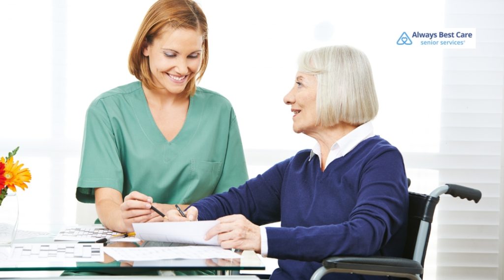 A caregiver in green scrubs is smiling and helping a senior woman in a wheelchair. The senior looks at the caregiver with a warm, appreciative expression while filling out paperwork together.