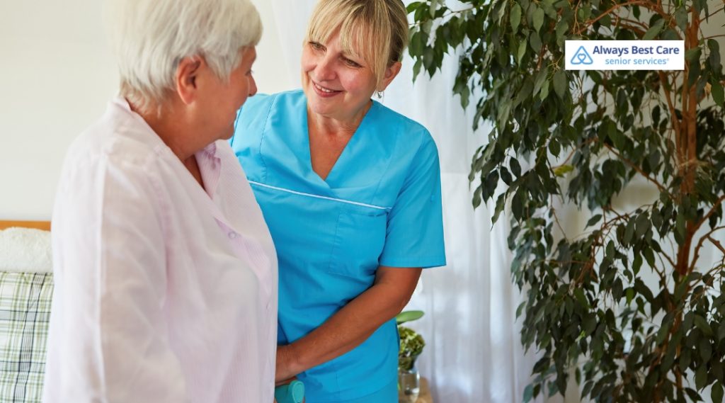 A caregiver in blue scrubs is assisting a senior woman in a bedroom, helping her with mobility. Both are smiling and having a pleasant interaction.