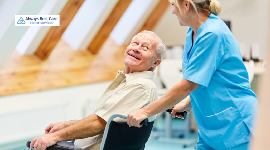 A senior man, seated in a wheelchair, is smiling up at his caregiver, who is pushing him in a bright, airy healthcare facility. Both seem engaged in a friendly and caring interaction.