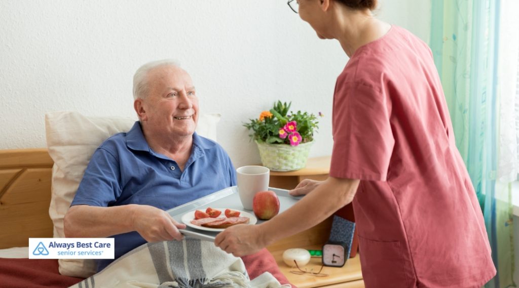 A caregiver in red scrubs is bringing a tray with breakfast to a senior man in bed. The senior man, smiling, is sitting up in bed, looking pleased as the caregiver presents the meal. The room is cozy, with bright plants and other homey touches around the bed.