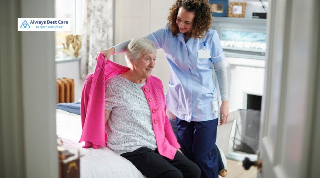 A caregiver assists a senior woman in a bedroom while helping her put on a pink cardigan, with a warm, caring expression.