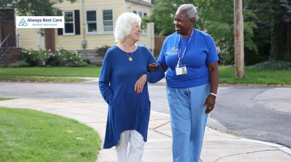 In-home caregiver walking arm in arm with a senior woman on a quiet neighborhood sidewalk, offering support and companionship.