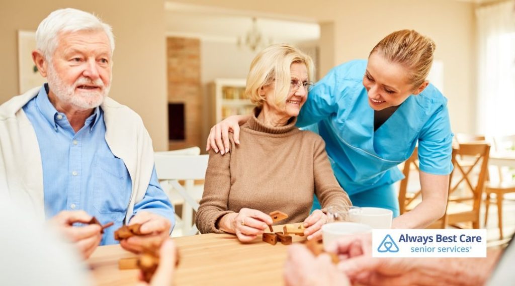 Caregiver assisting seniors during a tabletop activity at home, creating a supportive and engaging social moment.