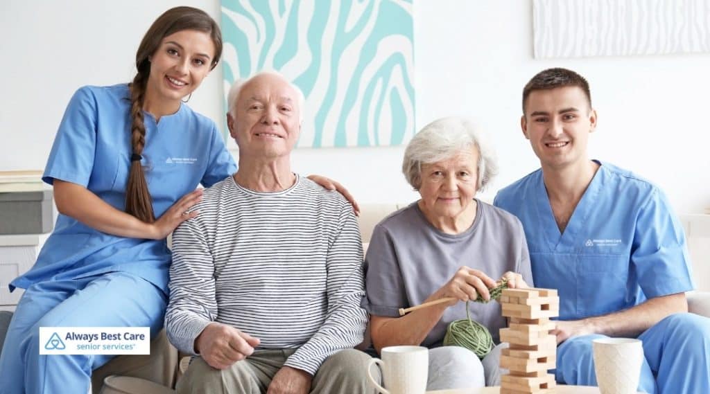 Two caregivers, one man and one woman in blue scrubs, are sitting with a senior couple, smiling together. The seniors are engaged in an activity of building a Jenga tower and knitting, surrounded by a calm and cozy living room space.