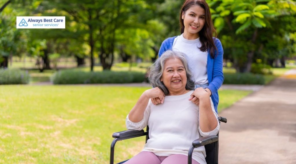 Caregiver pushing an older adult in a wheelchair along a park pathway