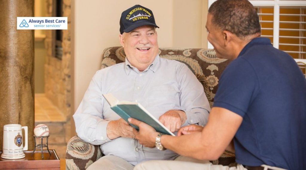 Care professional in blue uniform taking notes while talking with an older man seated on a patterned sofa at home.