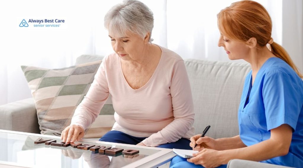 Senior woman sitting on a sofa arranging wooden letters that spell ‘dementia’ on a glass coffee table while a caregiver in blue scrubs takes notes beside her, Always Best Care logo in the corner.