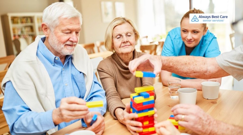 Group of seniors sitting around a table stacking bright wooden blocks together, with a caregiver in blue scrubs nearby and coffee mugs on the table, Always Best Care logo in the corner.
