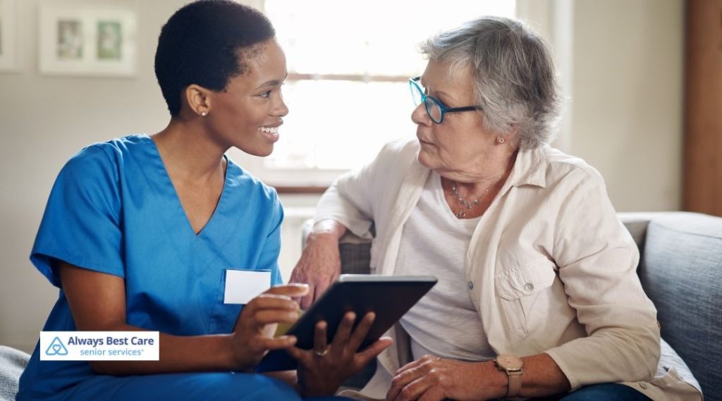 In-home caregiver in blue scrubs sitting on a couch, using a digital tablet to review care information with an older woman.