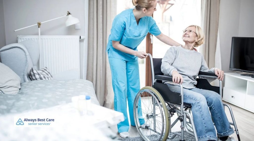 Caregiver standing beside an older woman seated in a wheelchair in her home.