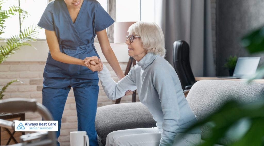 A caregiver in blue scrubs assists an elderly woman with getting up from the couch, offering her support as they engage in a helping and caring moment.