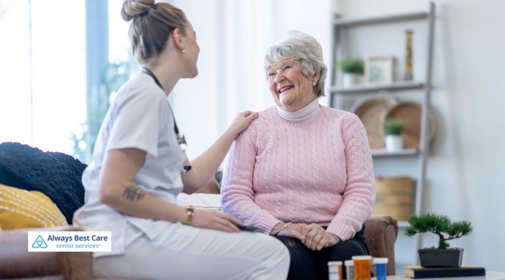 A caregiver in white scrubs smiles while offering comforting support to an elderly woman sitting on a couch, discussing medication or health matters as they share a pleasant conversation.