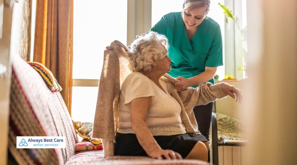 A caregiver in a green uniform helps an elderly woman by gently assisting her with putting on a sweater, ensuring her comfort and safety in the home.