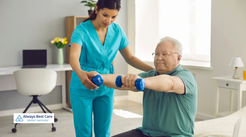 A caregiver in teal scrubs assists an elderly man with a light exercise using dumbbells, promoting health and well-being as part of his physical activity routine at home.