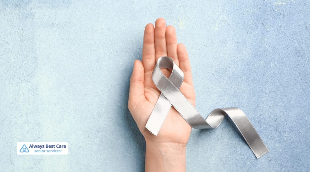 A silver awareness ribbon is held by an open hand against a light blue textured background, symbolizing advocacy for Parkinson’s disease awareness.